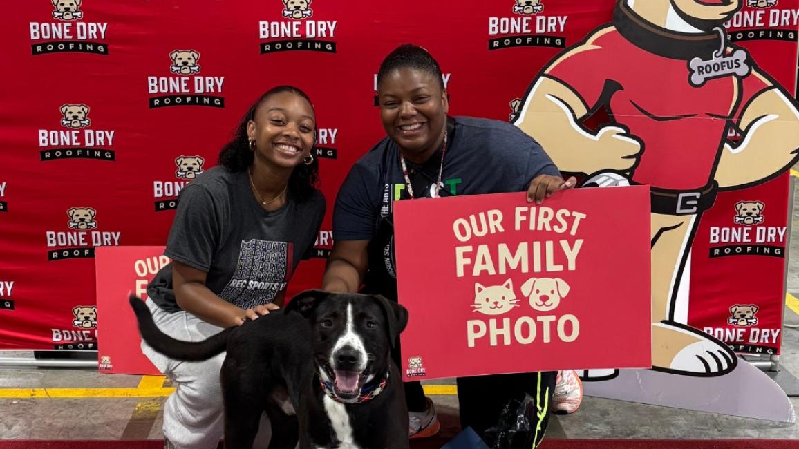 Two people with a dog holding a "Our first family photo" sign in front of a Bone Dry Roofing backdrop with a dog mascot