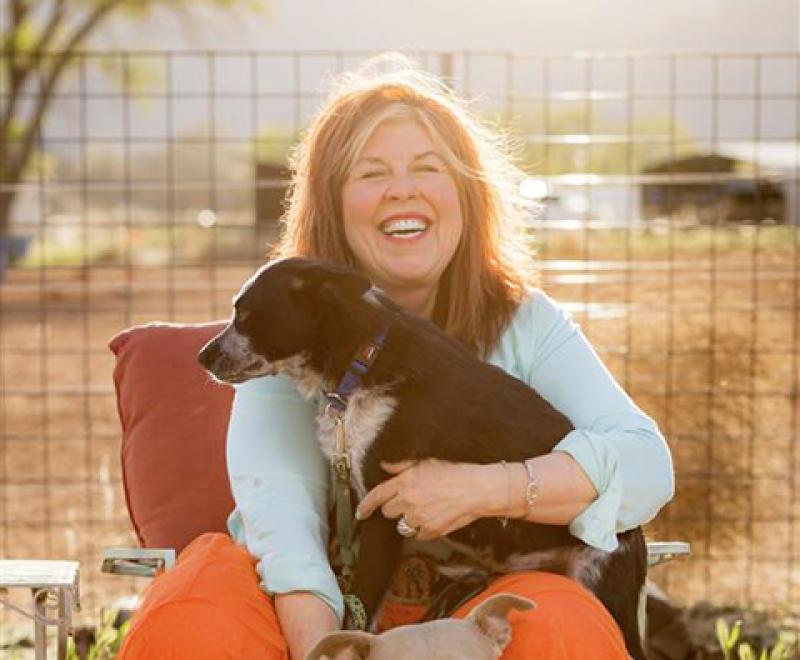 Patty Hegwood smiling and holding a small dog on her lap with a larger dog at her feet