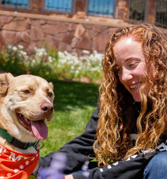 Woman with medium brown dog sitting in grass in front of Welcome Center